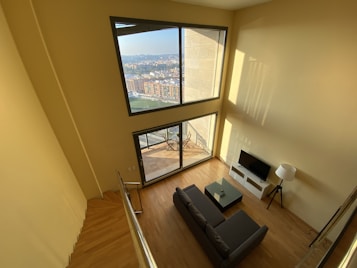 A spacious living room with large windows offering a panoramic city view. The interior features light wood flooring, a dark gray sofa, a small coffee table, a flat screen TV on a sleek white stand, and a floor lamp. The room is filled with natural light, enhancing the warm tones of the room.