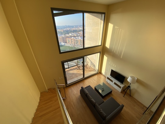 A spacious living room with large windows offering a panoramic city view. The interior features light wood flooring, a dark gray sofa, a small coffee table, a flat screen TV on a sleek white stand, and a floor lamp. The room is filled with natural light, enhancing the warm tones of the room.