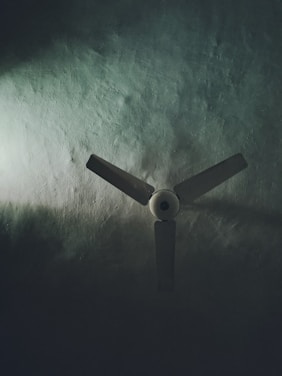 A three-blade ceiling fan set against a textured surface, partially illuminated by soft light emphasizing shadows