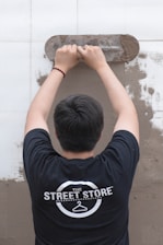 A person is viewed from behind while applying plaster to a wall using a trowel. The individual is wearing a black t-shirt with a logo that reads 'The Street Store'. The wall is partially covered in plaster, revealing a work in progress.