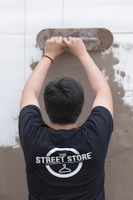 A person is viewed from behind while applying plaster to a wall using a trowel. The individual is wearing a black t-shirt with a logo that reads 'The Street Store'. The wall is partially covered in plaster, revealing a work in progress.