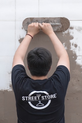 A person is viewed from behind while applying plaster to a wall using a trowel. The individual is wearing a black t-shirt with a logo that reads 'The Street Store'. The wall is partially covered in plaster, revealing a work in progress.