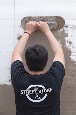 A person is viewed from behind while applying plaster to a wall using a trowel. The individual is wearing a black t-shirt with a logo that reads 'The Street Store'. The wall is partially covered in plaster, revealing a work in progress.
