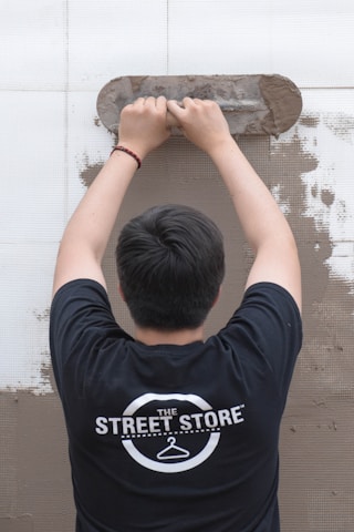 A person is viewed from behind while applying plaster to a wall using a trowel. The individual is wearing a black t-shirt with a logo that reads 'The Street Store'. The wall is partially covered in plaster, revealing a work in progress.