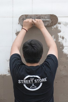 A person is viewed from behind while applying plaster to a wall using a trowel. The individual is wearing a black t-shirt with a logo that reads 'The Street Store'. The wall is partially covered in plaster, revealing a work in progress.