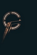 A circular arrangement of LED lights is mounted on a metal pole against a dark background. The lights form a partial circle with a gap on one side, illuminating the surrounding area with a warm glow.