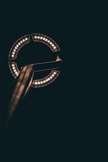 A circular arrangement of LED lights is mounted on a metal pole against a dark background. The lights form a partial circle with a gap on one side, illuminating the surrounding area with a warm glow.
