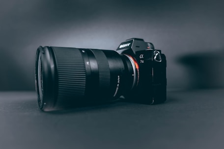 A professional camera with a large zoom lens is positioned at a slight angle, set against a smooth, dark, matte background. The camera and lens are prominently displayed, with the brand name visible on the camera body.