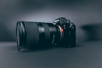 A professional camera with a large zoom lens is positioned at a slight angle, set against a smooth, dark, matte background. The camera and lens are prominently displayed, with the brand name visible on the camera body.