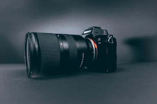 A professional camera with a large zoom lens is positioned at a slight angle, set against a smooth, dark, matte background. The camera and lens are prominently displayed, with the brand name visible on the camera body.