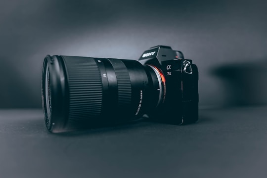 A professional camera with a large zoom lens is positioned at a slight angle, set against a smooth, dark, matte background. The camera and lens are prominently displayed, with the brand name visible on the camera body.