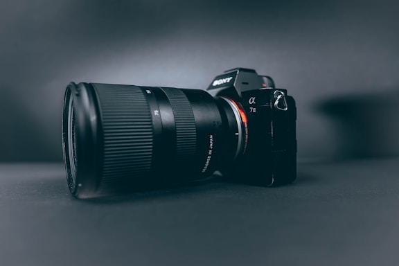 A professional camera with a large zoom lens is positioned at a slight angle, set against a smooth, dark, matte background. The camera and lens are prominently displayed, with the brand name visible on the camera body.
