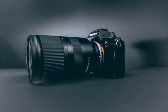 A professional camera with a large zoom lens is positioned at a slight angle, set against a smooth, dark, matte background. The camera and lens are prominently displayed, with the brand name visible on the camera body.