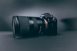 A professional camera with a large zoom lens is positioned at a slight angle, set against a smooth, dark, matte background. The camera and lens are prominently displayed, with the brand name visible on the camera body.