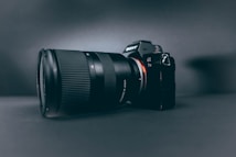 A professional camera with a large zoom lens is positioned at a slight angle, set against a smooth, dark, matte background. The camera and lens are prominently displayed, with the brand name visible on the camera body.