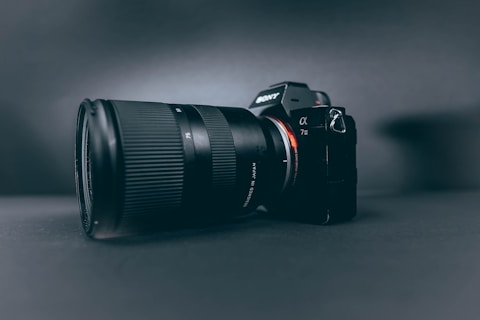 A professional camera with a large zoom lens is positioned at a slight angle, set against a smooth, dark, matte background. The camera and lens are prominently displayed, with the brand name visible on the camera body.