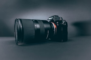 A professional camera with a large zoom lens is positioned at a slight angle, set against a smooth, dark, matte background. The camera and lens are prominently displayed, with the brand name visible on the camera body.