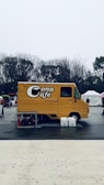 A yellow food truck labeled 'Cona Cafe' is parked in an open outdoor area. Leafless trees are visible in the background under a gray sky, and the truck is surrounded by a few objects like tables, chairs, and buckets.