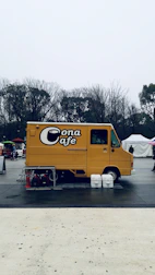 A yellow food truck labeled 'Cona Cafe' is parked in an open outdoor area. Leafless trees are visible in the background under a gray sky, and the truck is surrounded by a few objects like tables, chairs, and buckets.