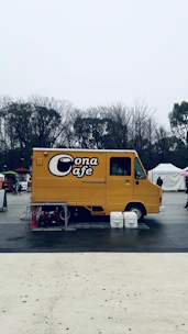 A yellow food truck labeled 'Cona Cafe' is parked in an open outdoor area. Leafless trees are visible in the background under a gray sky, and the truck is surrounded by a few objects like tables, chairs, and buckets.