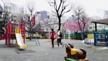 A playground with colorful equipment, including a yellow slide and climbing structures, is surrounded by bare trees and a blossoming pink cherry blossom tree. In the background are urban buildings. Two people are walking through the playground, carrying backpacks. A ride-on spring animal toy is visible in the foreground.