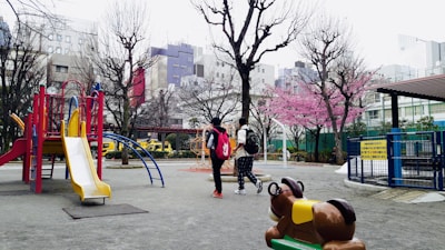 A playground with colorful equipment, including a yellow slide and climbing structures, is surrounded by bare trees and a blossoming pink cherry blossom tree. In the background are urban buildings. Two people are walking through the playground, carrying backpacks. A ride-on spring animal toy is visible in the foreground.