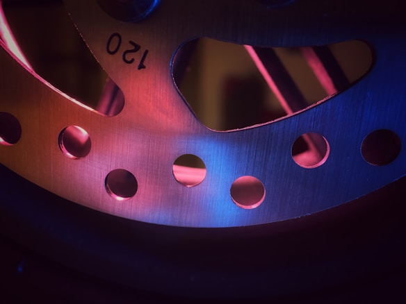 A close-up view of a metal surface with circular perforations arranged in a symmetrical pattern. The surface is illuminated with a combination of pink and blue lighting, creating a gradient effect. The number 120 is visible, suggesting a measurement or component specification. The overall texture appears brushed and metallic.