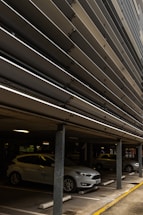 A multi-story parking structure with several cars parked, including a white car in the foreground. Metal slats are visible on the upper levels, providing a modern architectural element. The parking space is marked with numbers, and there are concrete pillars supporting the ceiling.