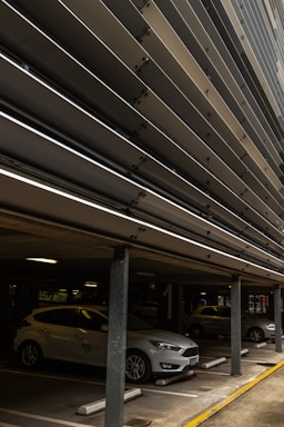 A multi-story parking structure with several cars parked, including a white car in the foreground. Metal slats are visible on the upper levels, providing a modern architectural element. The parking space is marked with numbers, and there are concrete pillars supporting the ceiling.