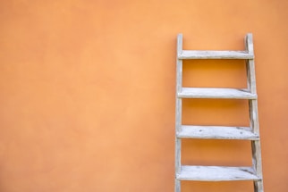 A wooden ladder is placed against a smooth, orange wall. The wall has a subtle texture, and the ladder has a rustic appearance with visible wood grain.