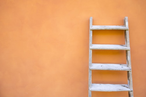 A wooden ladder is placed against a smooth, orange wall. The wall has a subtle texture, and the ladder has a rustic appearance with visible wood grain.
