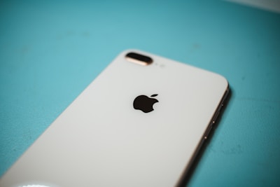A close-up of a white smartphone with a prominent Apple logo, placed on a turquoise surface. The camera and flash are visible on the top corner of the phone.