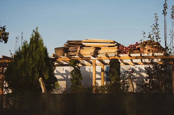 A stack of wooden planks and orange pipes is neatly arranged on a rooftop structure, surrounded by green plants and trees. The structure is made of wooden beams and has a white wall visible in the background. The sky is clear and blue, giving the scene ample natural light.