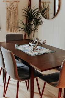 A cozy dining room with a dark wooden table centerpiece adorned with decorative antlers and a cloth. Surrounding the table are gray upholstered chairs with wooden legs. A potted plant and bohemian-style macrame wall hangings add a touch of nature and texture. A round mirror is mounted on the wall, reflecting part of the room.