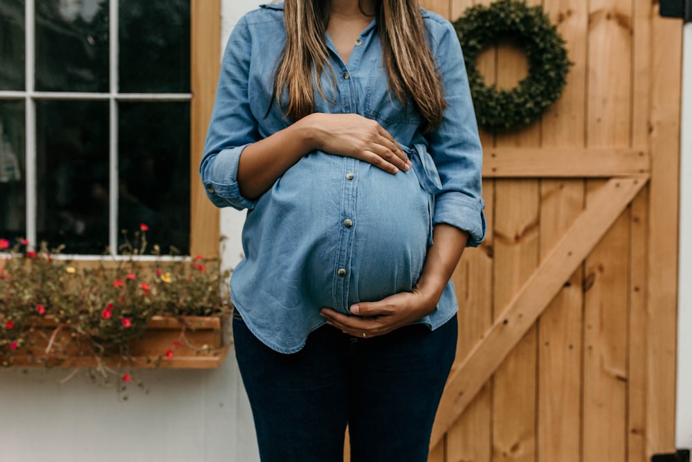 woman in blue denim button up jacket and blue denim jeans standing near brown wooden fence