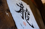A piece of textured paper with bold black calligraphy characters displayed on a wooden surface. A red seal or stamp is present near the bottom of the paper, adding an artistic touch.