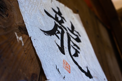 A piece of textured paper with bold black calligraphy characters displayed on a wooden surface. A red seal or stamp is present near the bottom of the paper, adding an artistic touch.