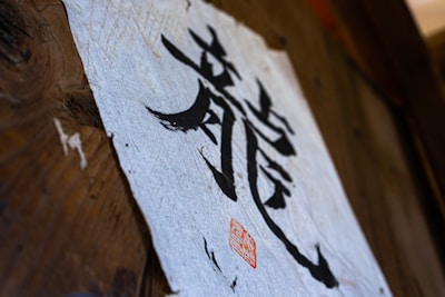 A piece of textured paper with bold black calligraphy characters displayed on a wooden surface. A red seal or stamp is present near the bottom of the paper, adding an artistic touch.