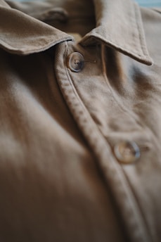 A close-up view of a brown fabric shirt focusing on the buttons and collar. The texture of the fabric is visible, showing the stitching details and smooth surface.