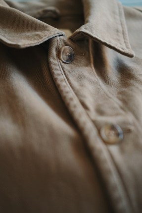 A close-up view of a brown fabric shirt focusing on the buttons and collar. The texture of the fabric is visible, showing the stitching details and smooth surface.