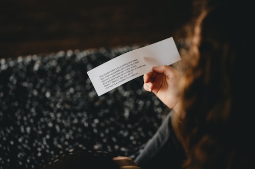 A person is holding a piece of paper with printed text. The text is partially visible and appears to resemble a message or poem. The background is blurred, predominantly dark with some scattered white dots.