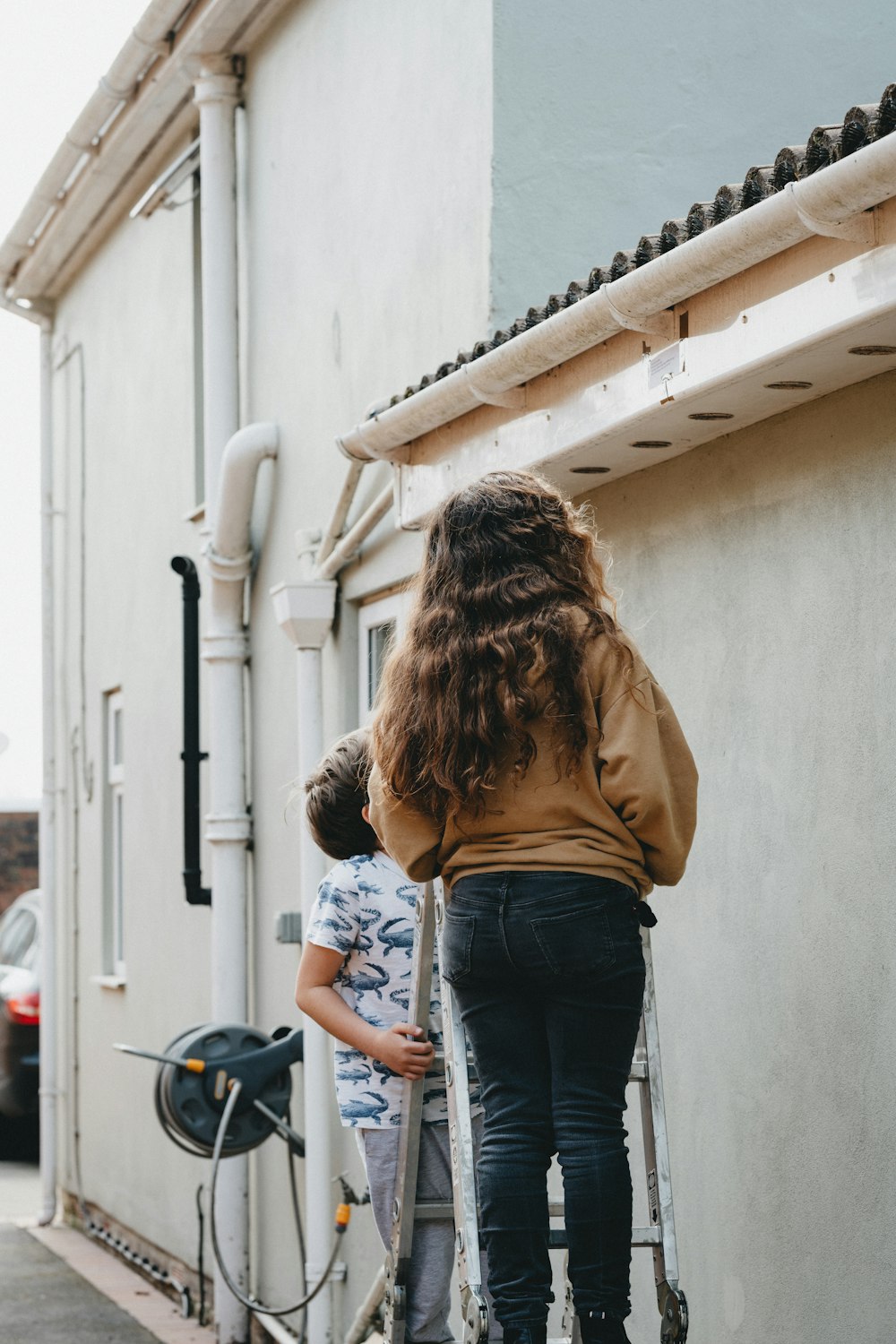 Femme en veste marron et jean bleu debout près du mur blanc pendant la journée