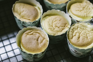 Several light brown, fluffy pastries are sitting in decorative paper cups resting on a black cooling rack. The pastries have a soft, slightly wrinkled surface, suggesting they are freshly baked and possibly cheesecakes or sponge cakes.