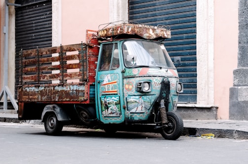 A colorful and well-worn three-wheeled vehicle, likely a small truck or tuk-tuk, is parked on a street. The vehicle features intricate, artistic designs and illustrations on its cabin and sides, including various vibrant patterns and images. It is situated against an urban backdrop with closed metal shutters.