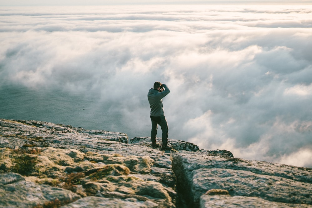 man in black jacket and black pants standing on rock formation near sea of clouds