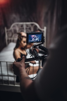 A camera operator is filming a woman who is sitting on a bed with an intricate metal headboard. The woman appears to be the focus of the video shoot, as she is visible on the camera's flip-out screen. The scene seems to have a dim, moody lighting setup.
