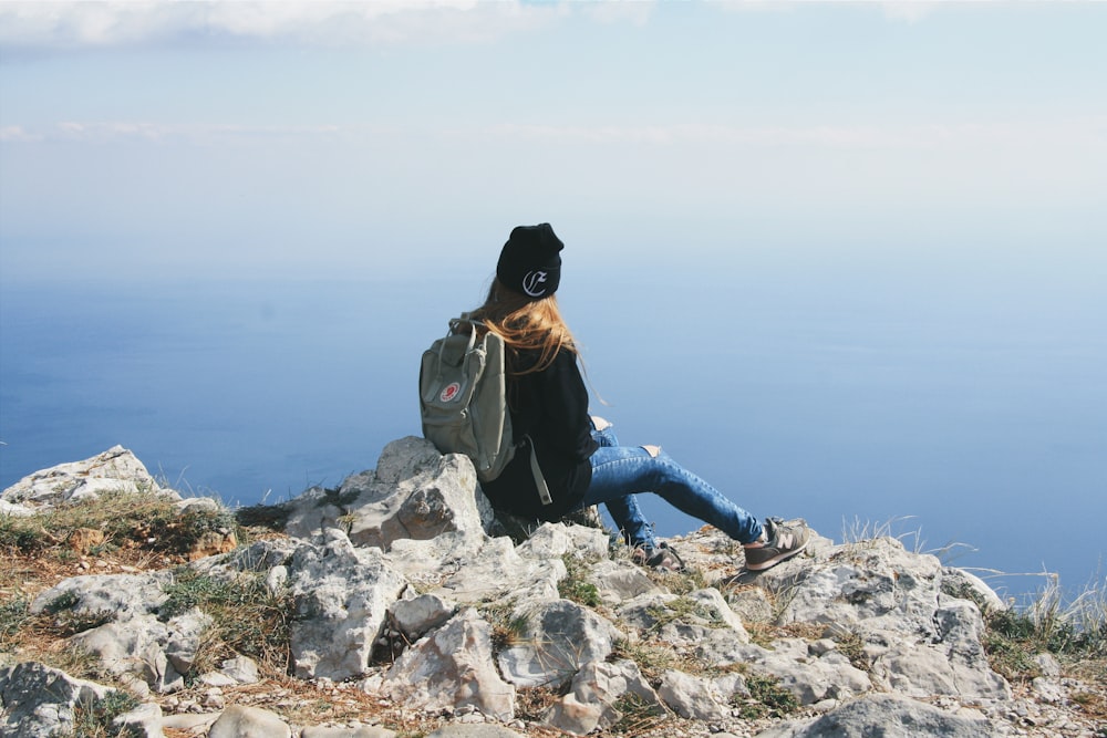 person in blue denim jeans and black jacket sitting on rock formation near body of water