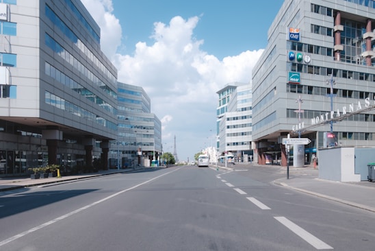 A wide urban street flanked by modern office buildings, under a bright blue sky with fluffy clouds. The setting is relatively empty with sparse traffic and a prominent sign in the foreground indicating location information.