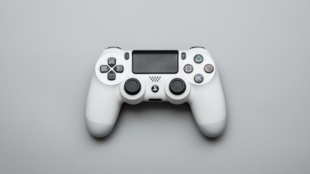 A white gaming controller is positioned on a light gray surface. The controller features dual analog sticks, a directional pad, and iconic four-button layout with distinct symbols. The design is sleek and modern, with additional features such as triggers and a central touchpad.