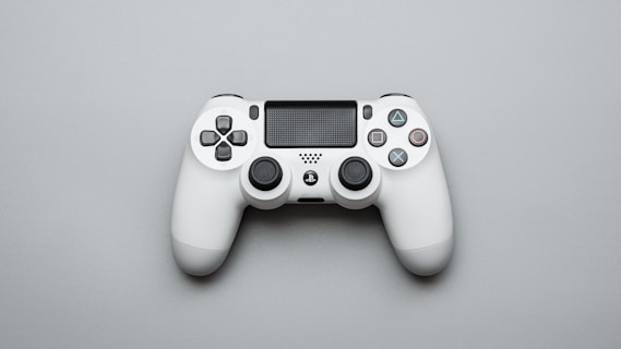 A white gaming controller is positioned on a light gray surface. The controller features dual analog sticks, a directional pad, and iconic four-button layout with distinct symbols. The design is sleek and modern, with additional features such as triggers and a central touchpad.